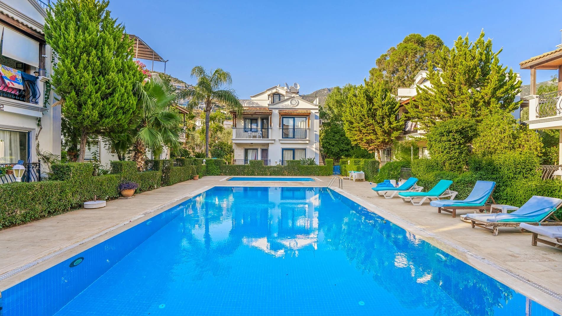 Ağaçlar ve şezlonglarla çevrili açık havuz ve arka planda villa