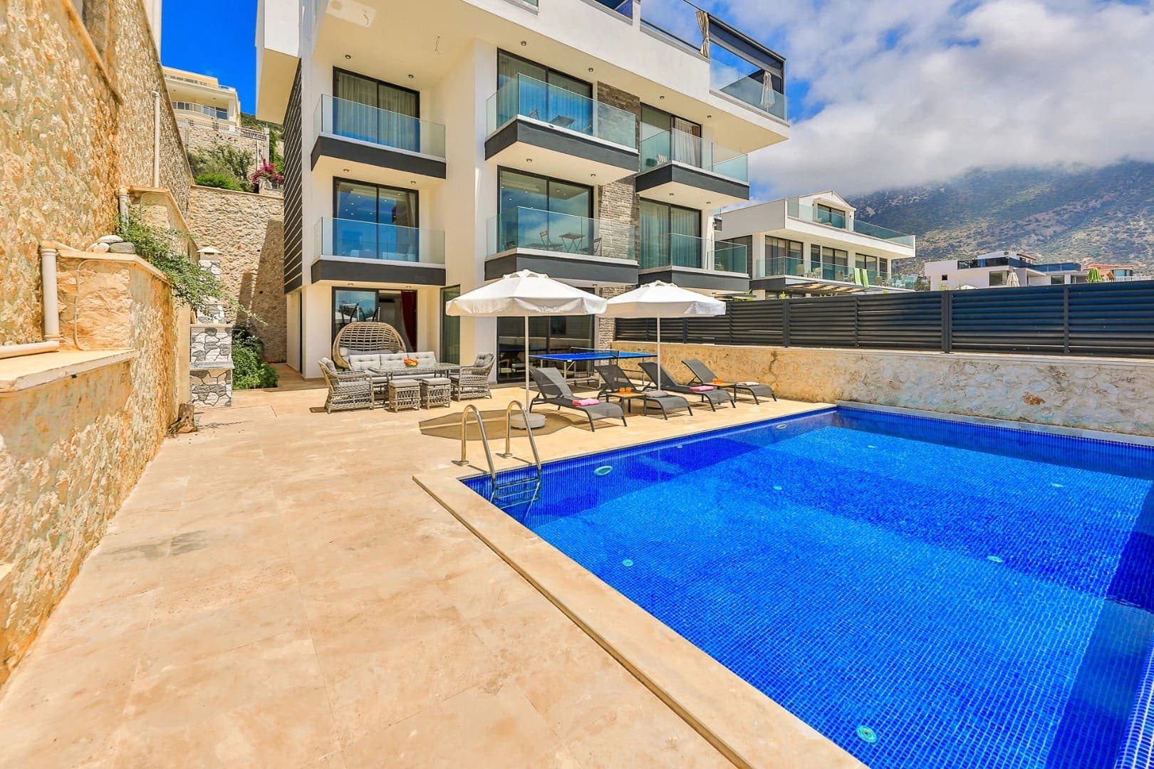 Beyaz çok katlı villa, önünde şezlonglar, güneş şemsiyeleri ve mavi yüzme havuzu.