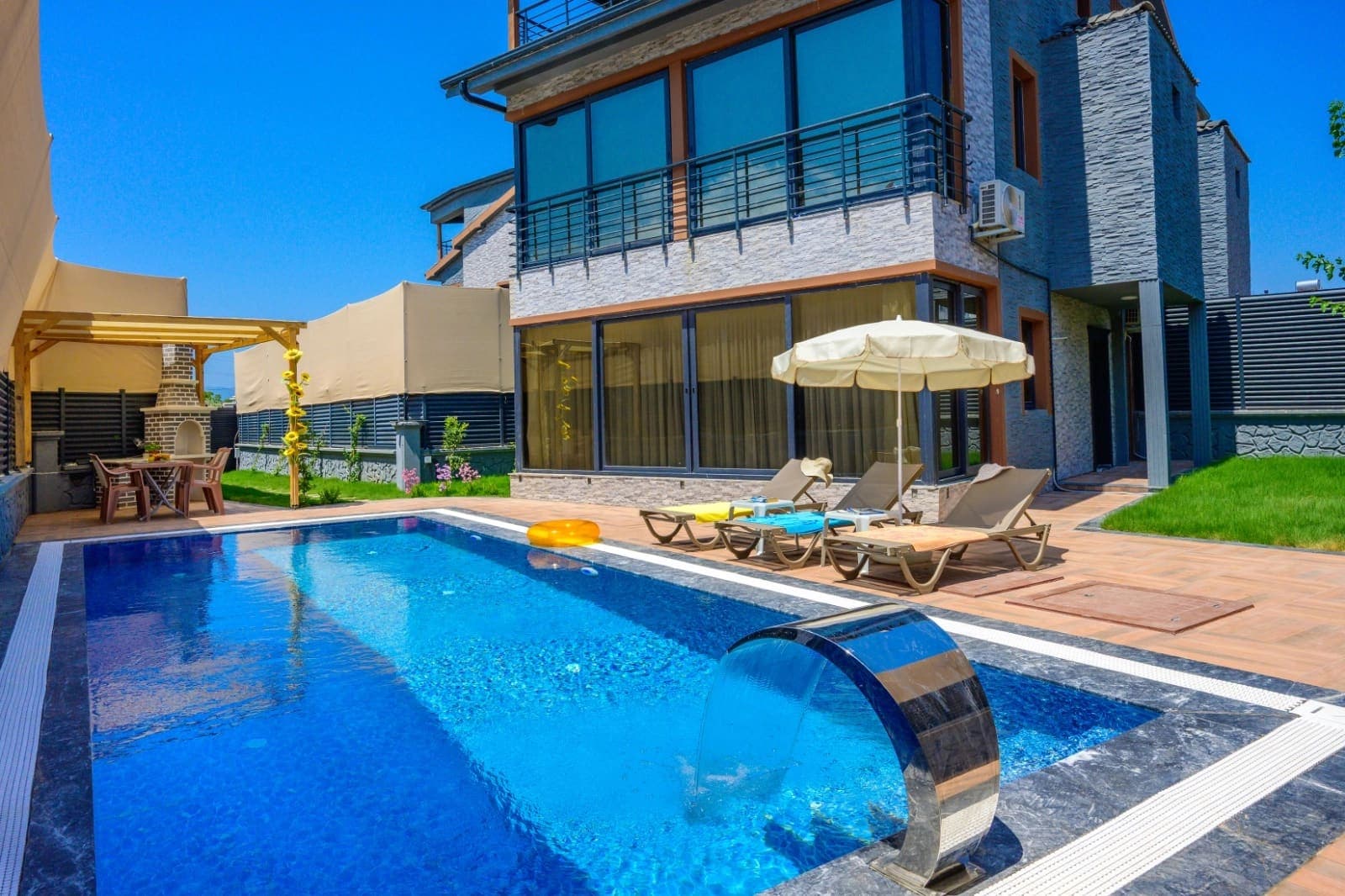 Mavi yüzme havuzu ve şezlongların bulunduğu taş cepheli villa bahçesi.