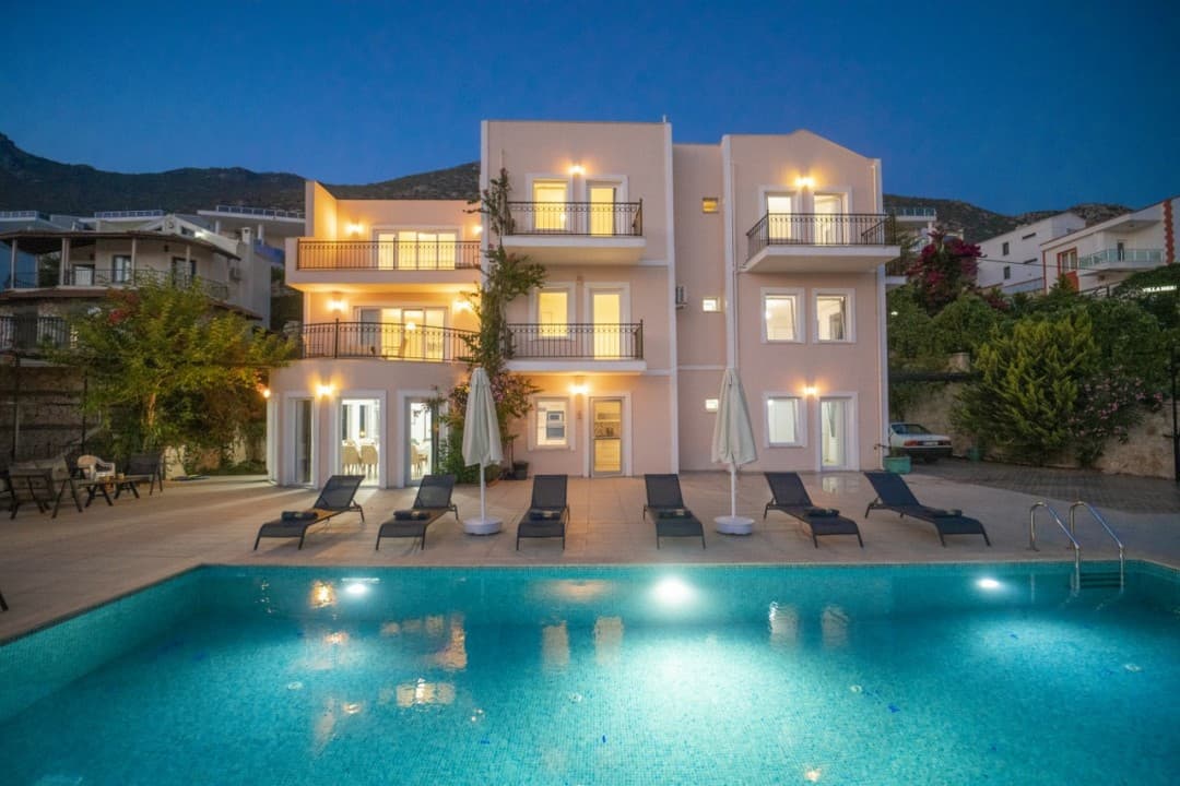 Akşam vakti aydınlatılmış havuzun başında şezlonglar ve arka planda çok katlı villa yer alıyor.