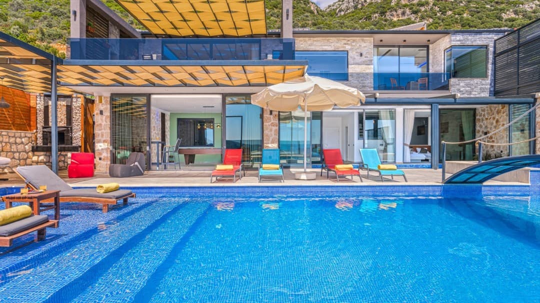 Gündüz vakti havuz kenarında şezlonglar, şemsiye ve arkada taş ve cam detaylı villa.