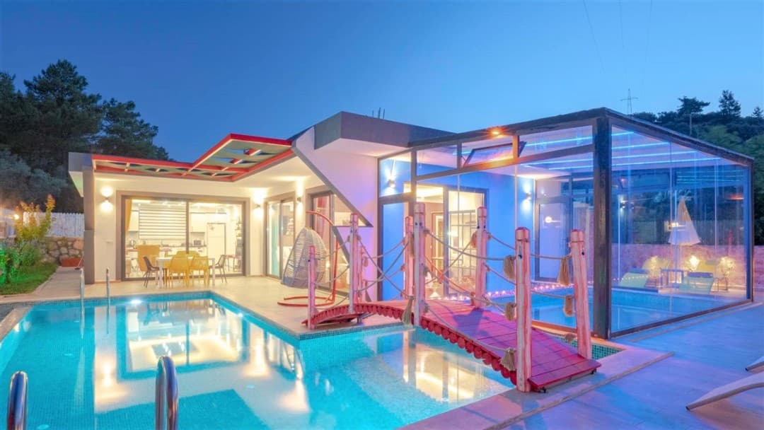 Akşamüstü aydınlatmalı havuz, kırmızı köprü ve cam kaplı kapalı havuzlu modern villa.