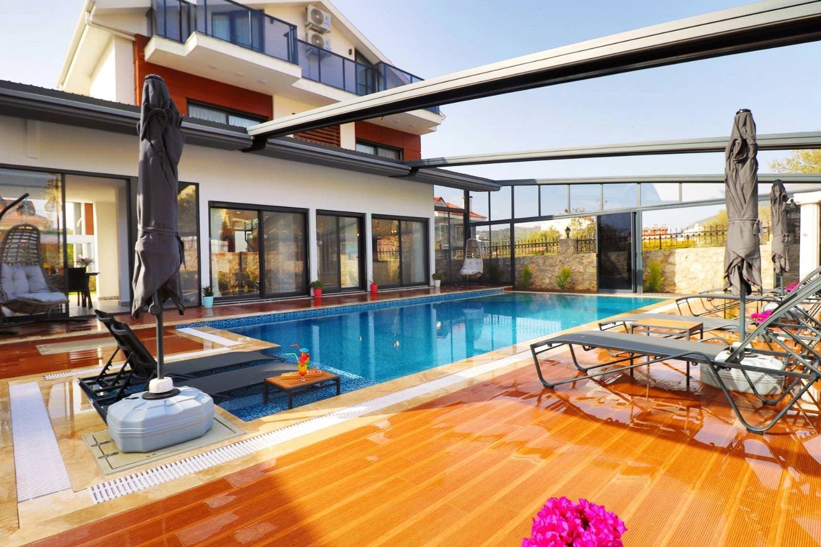 Açılır kapanır tavanlı havuz ve çevresinde şezlonglar bulunan modern villa bahçesi.