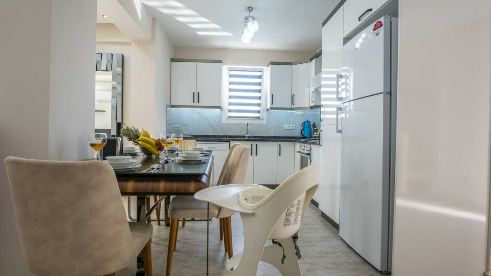 Beyaz dolaplı mutfakta yemek masası ve ön planda beyaz mama sandalyesi var.