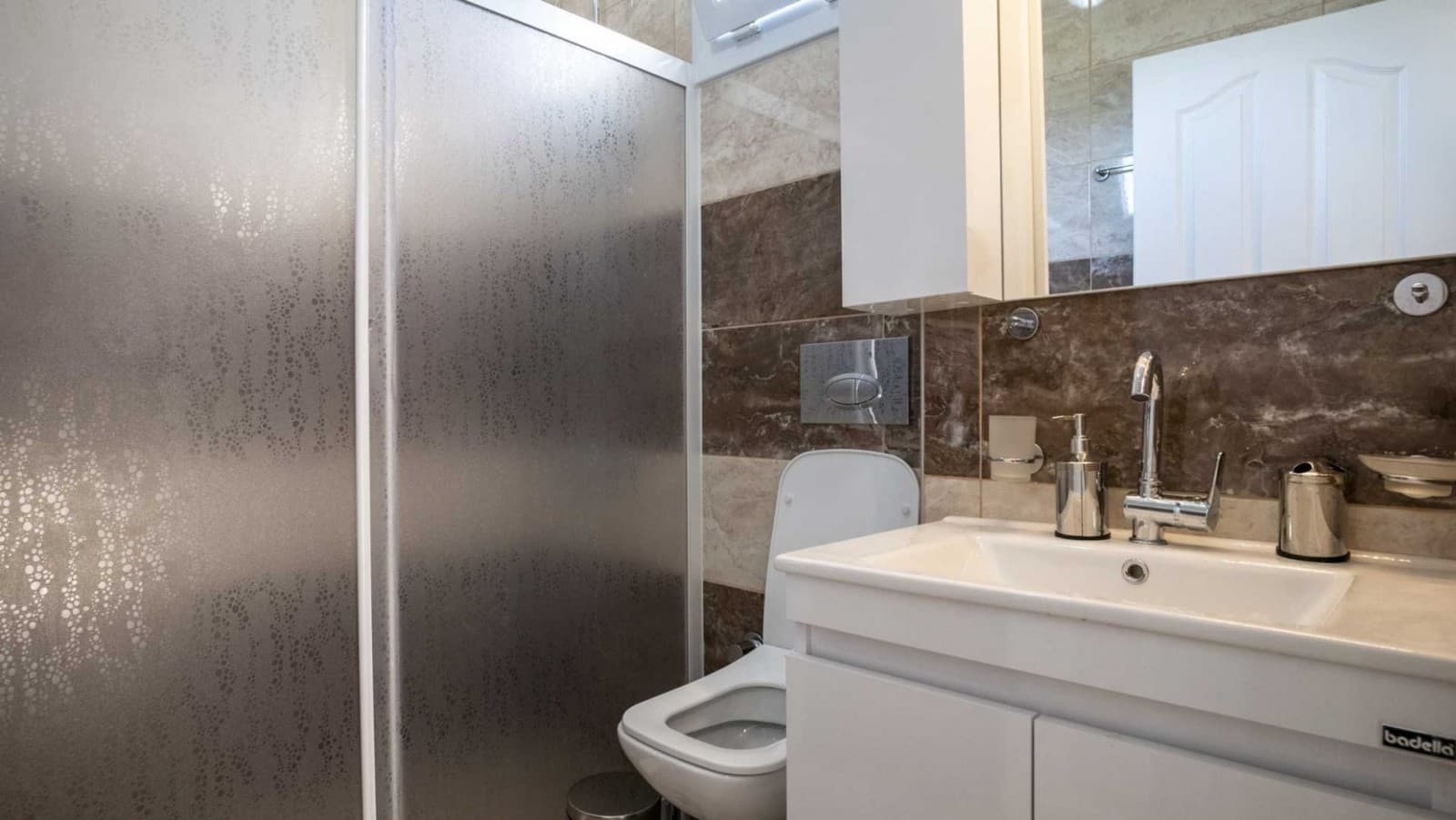 Kahverengi mermer detaylı banyoda beyaz lavabo, klozet ve duşakabin yer alıyor.