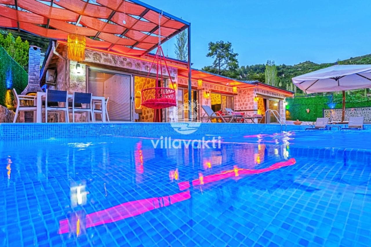 Akşam vakti aydınlatılmış taş villa, havuz kenarında şezlonglar ve kırmızı asma salıncak.