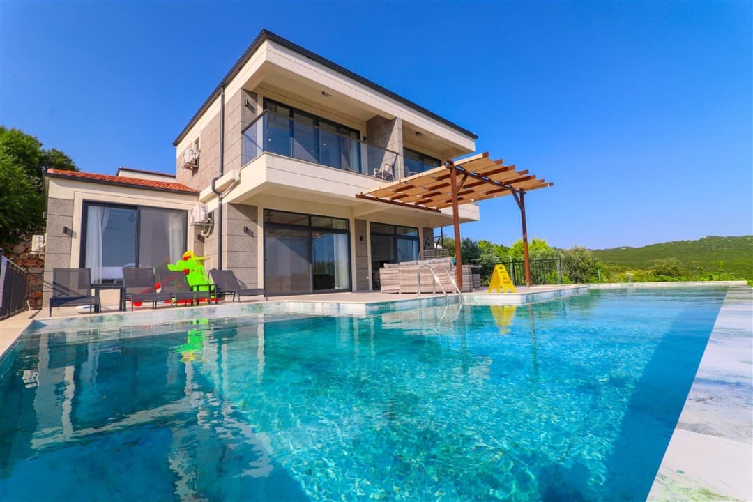Önünde sonsuzluk havuzu, şezlonglar ve ahşap pergolalı oturma alanı bulunan iki katlı modern villa.