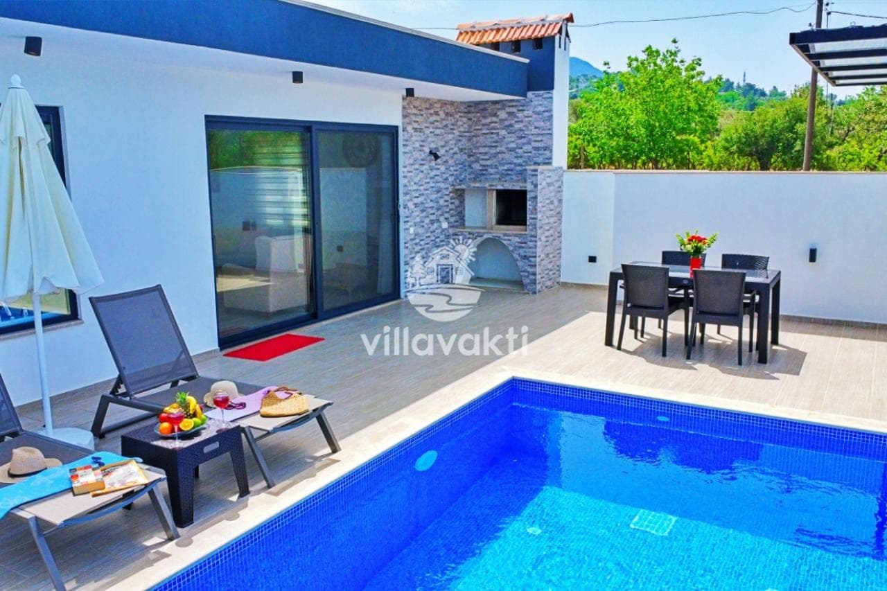 Havuz kenarında şezlonglar, yemek masası ve taş kaplamalı barbekü alanı bulunan villa terası.