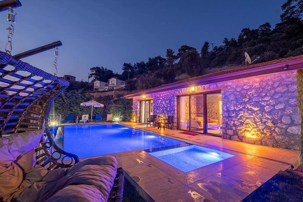 Gece aydınlatılmış taş duvarlı villa, havuz ve asma salıncak içeren dış mekan.