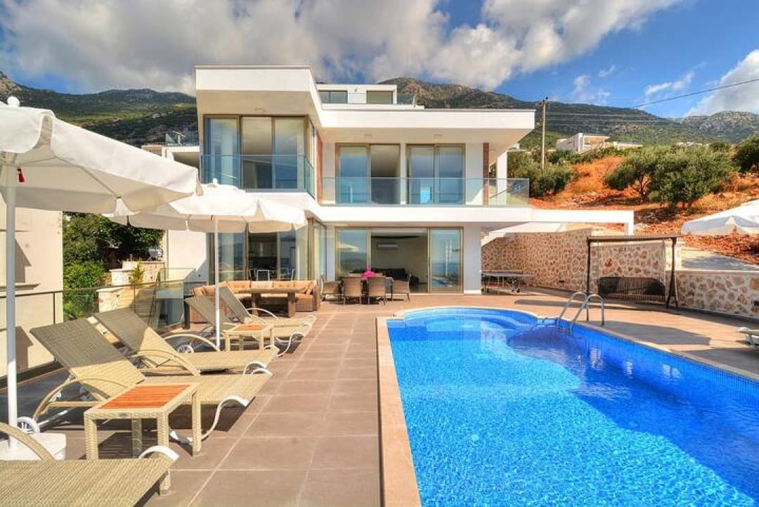 Dağ manzaralı, havuz başında şezlonglar ve beyaz şemsiyeler bulunan iki katlı modern villa.