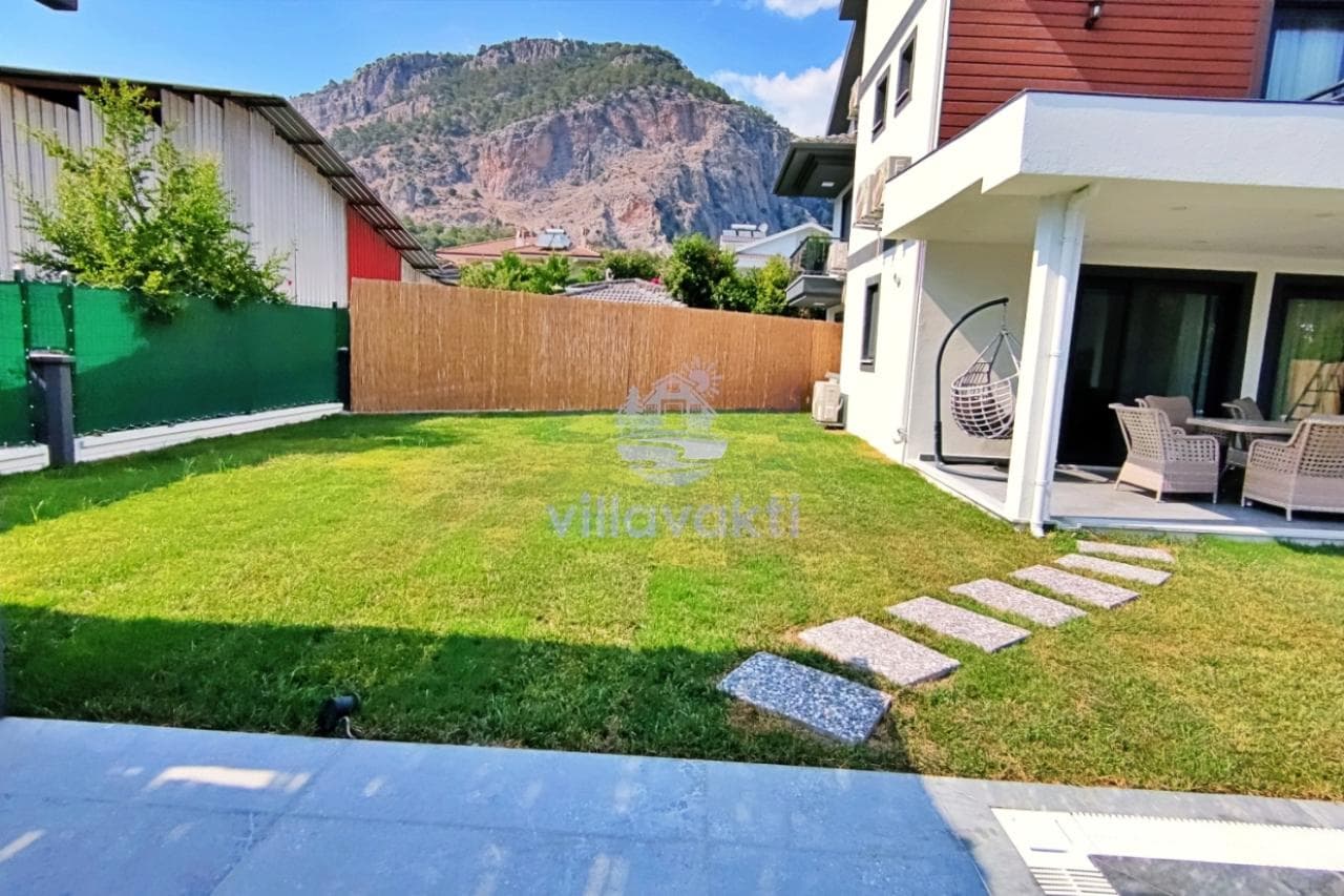 Villa bahçesinde yeşil çim alan, taş zeminli veranda, asılı koltuk ve dağ manzarası.