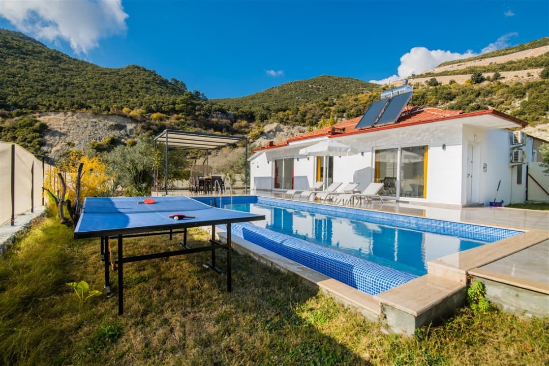 Dağ manzaralı, bahçesinde masa tenisi ve yüzme havuzu bulunan beyaz dış cepheli tek katlı villa.