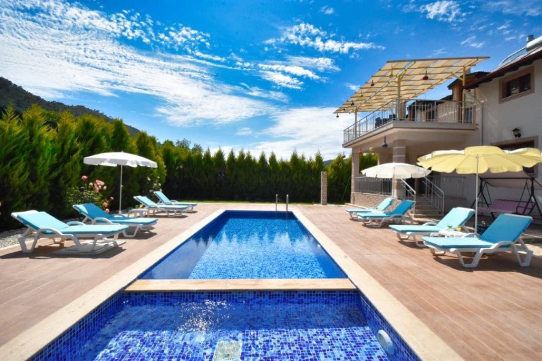 Mavi havuzlu, şezlonglu ve güneş şemsiyeli villa bahçesi, arkada ağaçlar ve açık mavi gökyüzü.