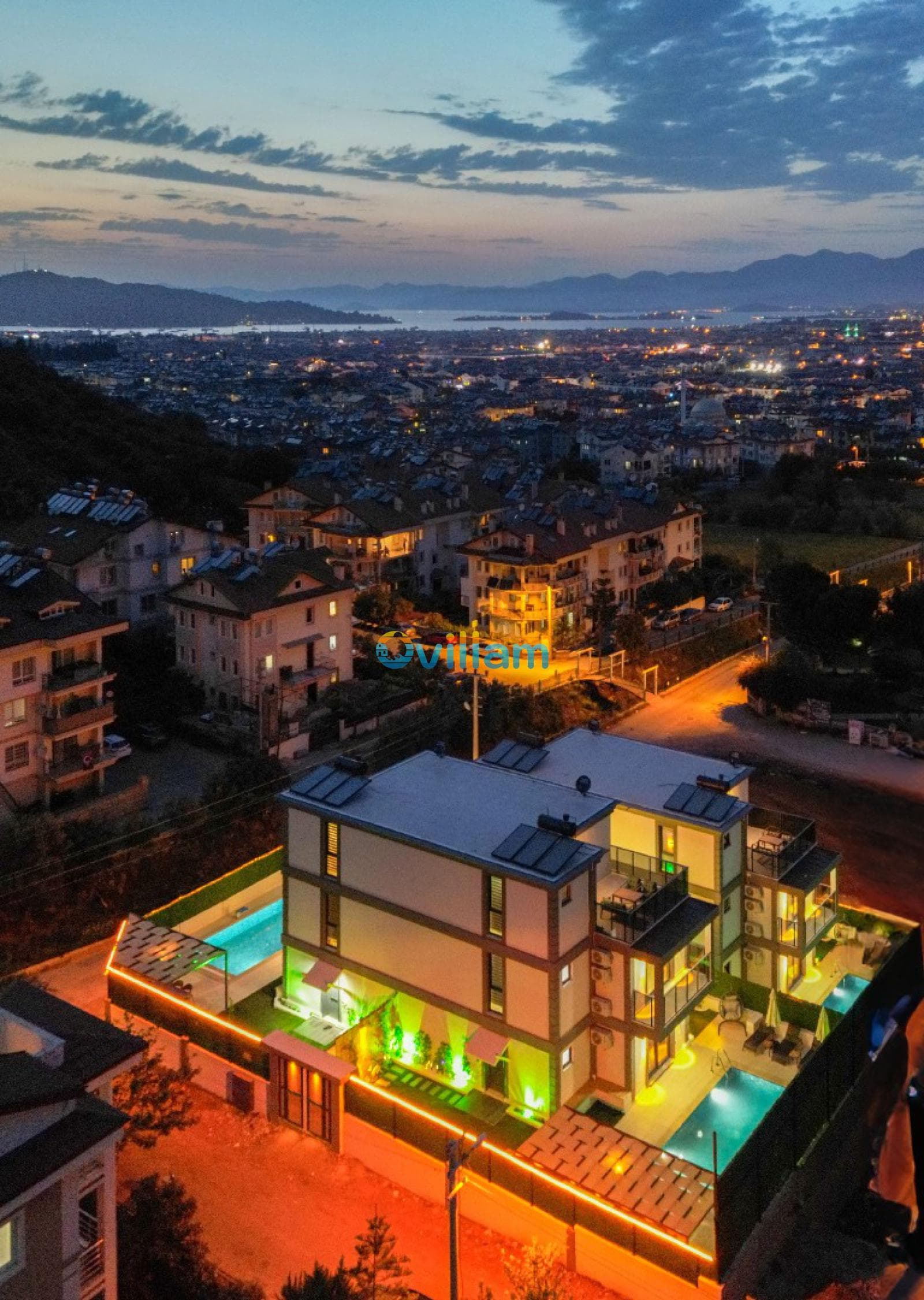 Gece ışıklandırılmış villa, havuzlar ve arkada şehir ile deniz manzarası.