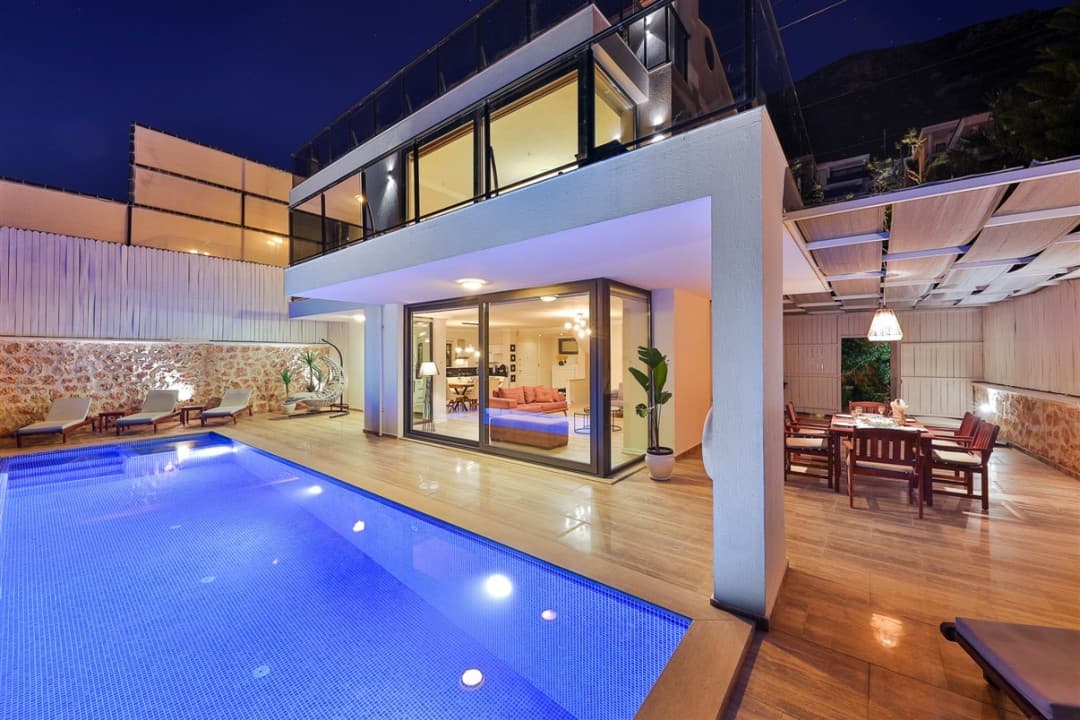Aydınlatmalı havuzu ve yemek masası olan iki katlı modern villanın akşam dış cephesi.