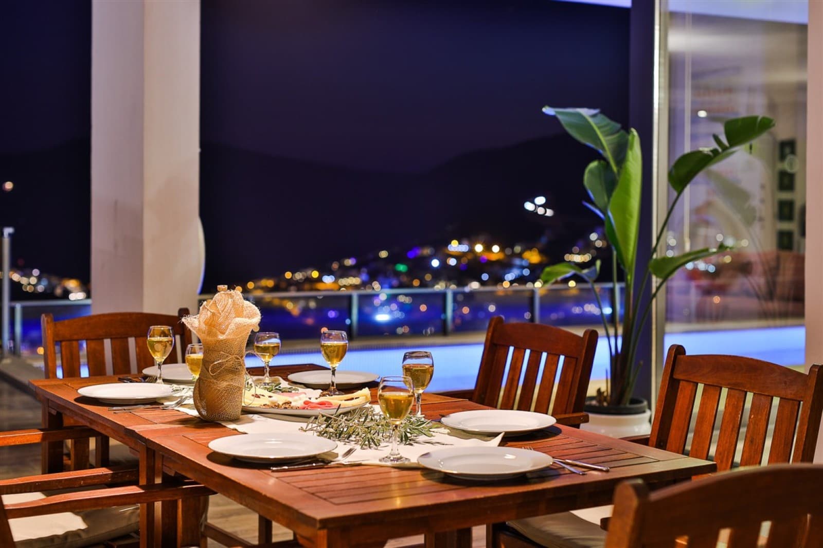Gece vakti ışıklandırılmış havuz manzaralı ahşap yemek masası ve şarap kadehleri.