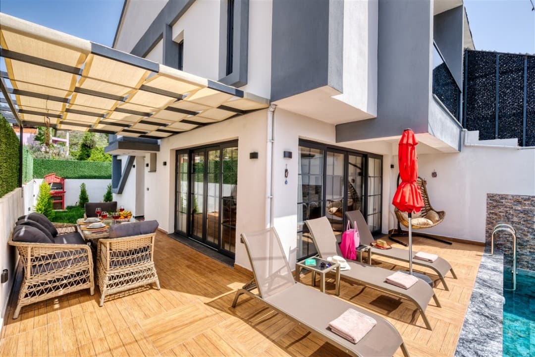Ahşap zeminli terasta hasır oturma grubu, şezlonglar, kırmızı şemsiye ve havuz kenarı bulunan modern villa.