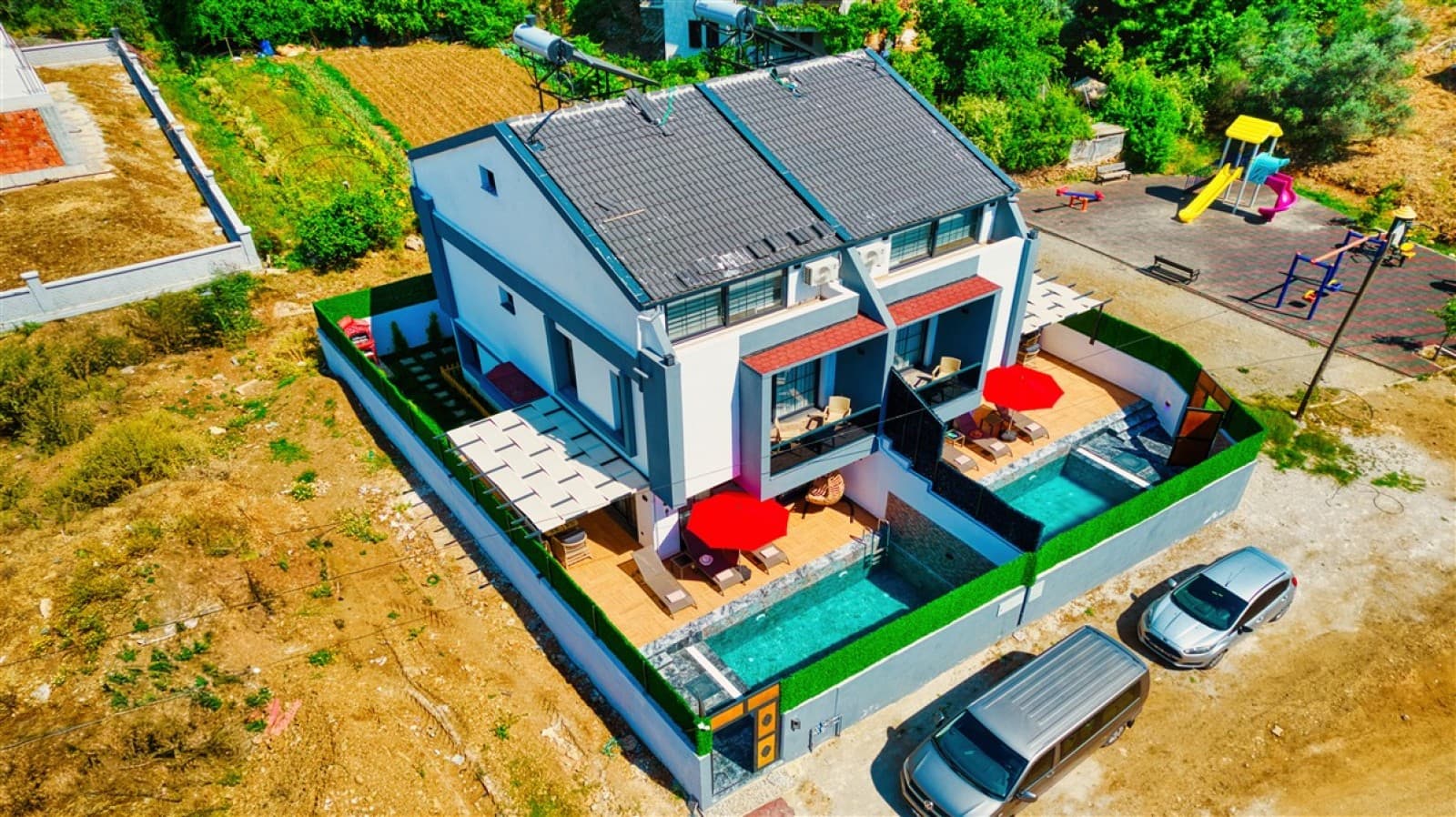Havuzlu, iki kırmızı şemsiyeli ve çocuk parkı yanındaki villanın havadan görünümü.