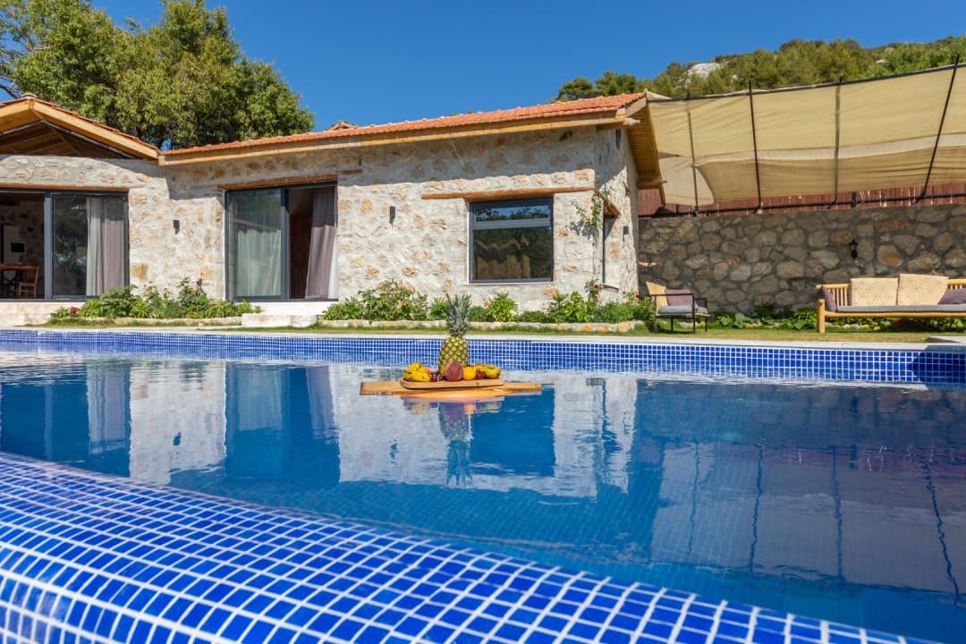 Mavi havuzun üstünde meyve tepsisi olan, taş duvarlı ve kiremit çatılı tek katlı villa.