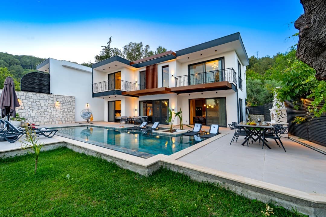 Önünde yüzme havuzu, şezlonglar ve yemek masası bulunan, iki katlı, modern mimariye sahip beyaz villa.