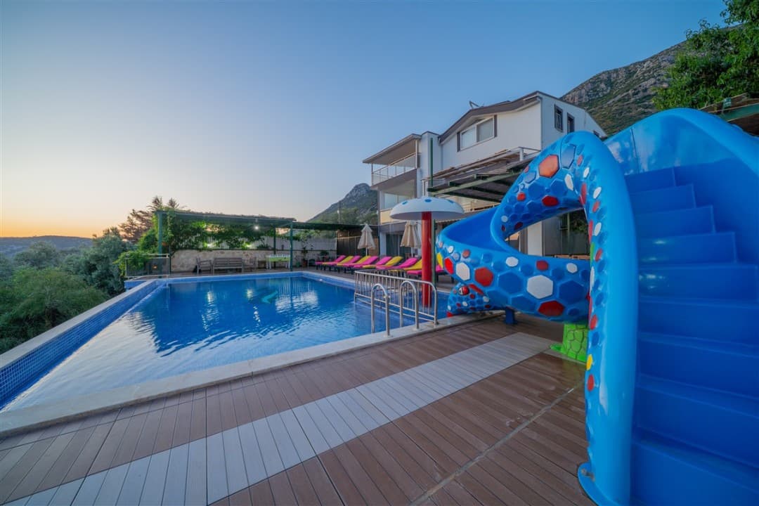 Mavi su kaydıraklı yüzme havuzu, şezlonglar ve gün batımında dağ manzaralı iki katlı villa.