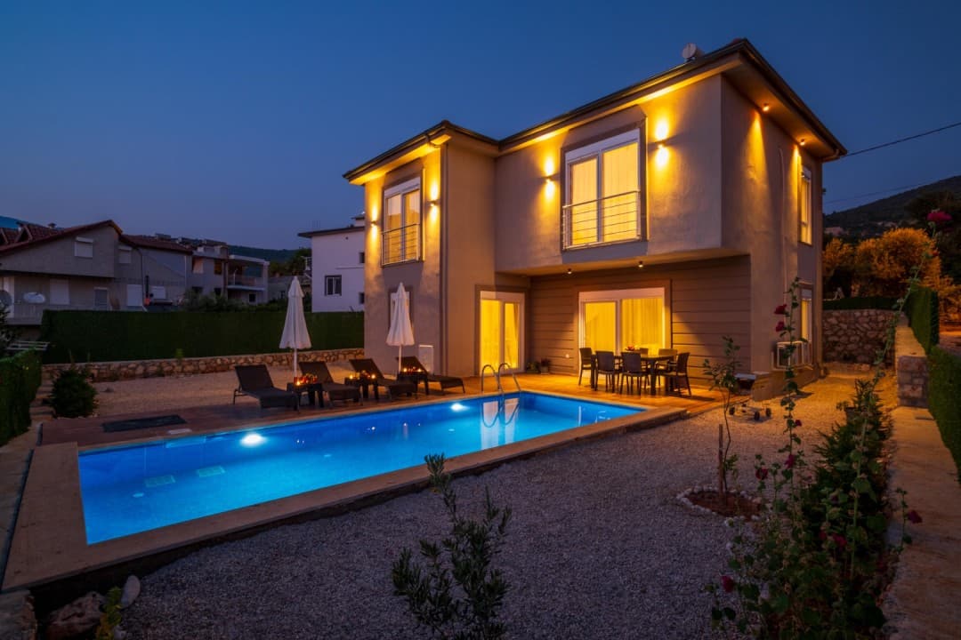 Akşam karanlığında dış cephesi aydınlatılmış iki katlı villa, havuzu ve şezlongları.
