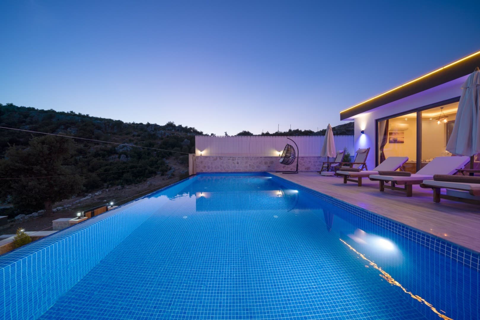 Gün batımında havuz kenarında şezlonglar ve tepe manzaralı modern villa.