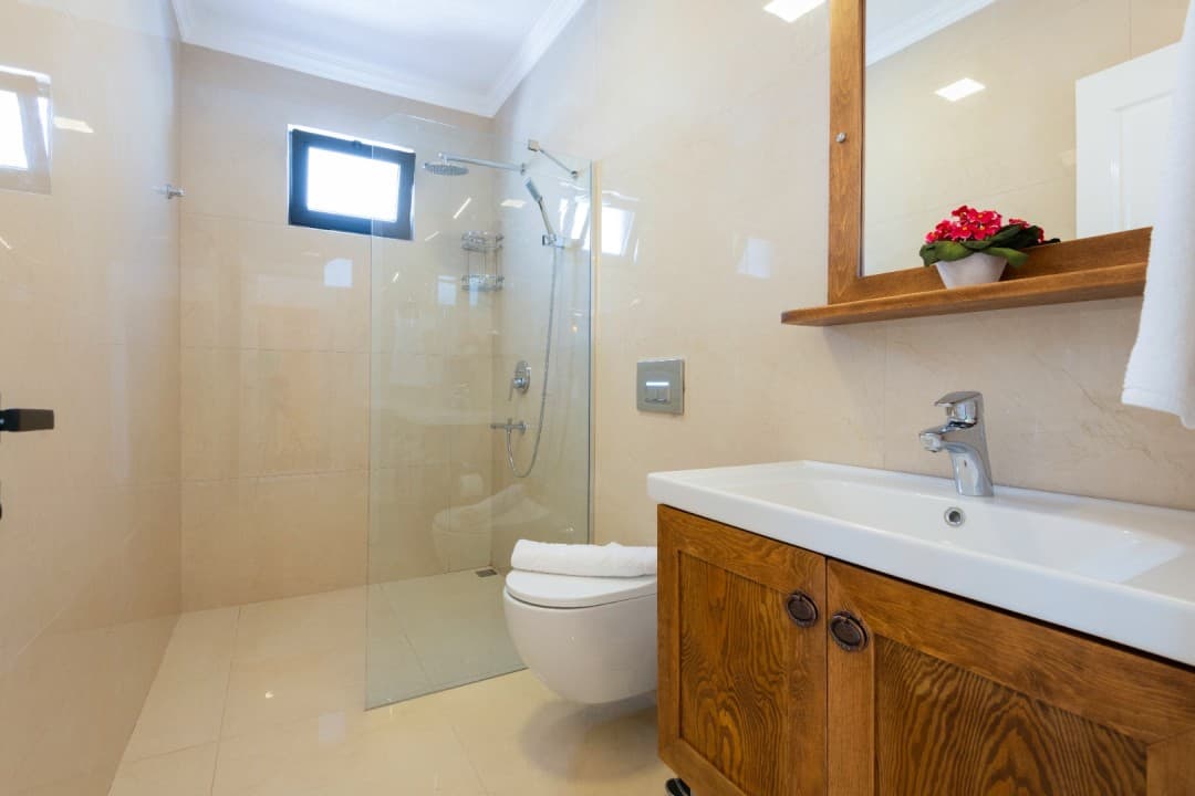 Bej tonlarındaki banyoda ahşap dolaplı lavabo ve cam duşakabin var.