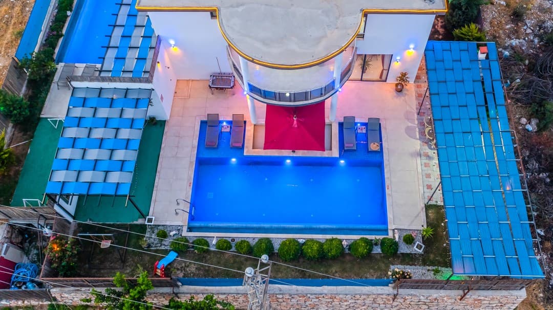 Havuzlu ve şezlonglu villanın havadan gece aydınlatmalı görünümü.