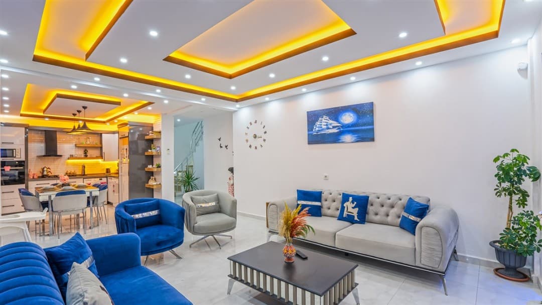 Sarı gizli aydınlatmalı tavanı olan, açık mutfaklı, mavi ve gri koltuk takımlı modern bir oturma odası.