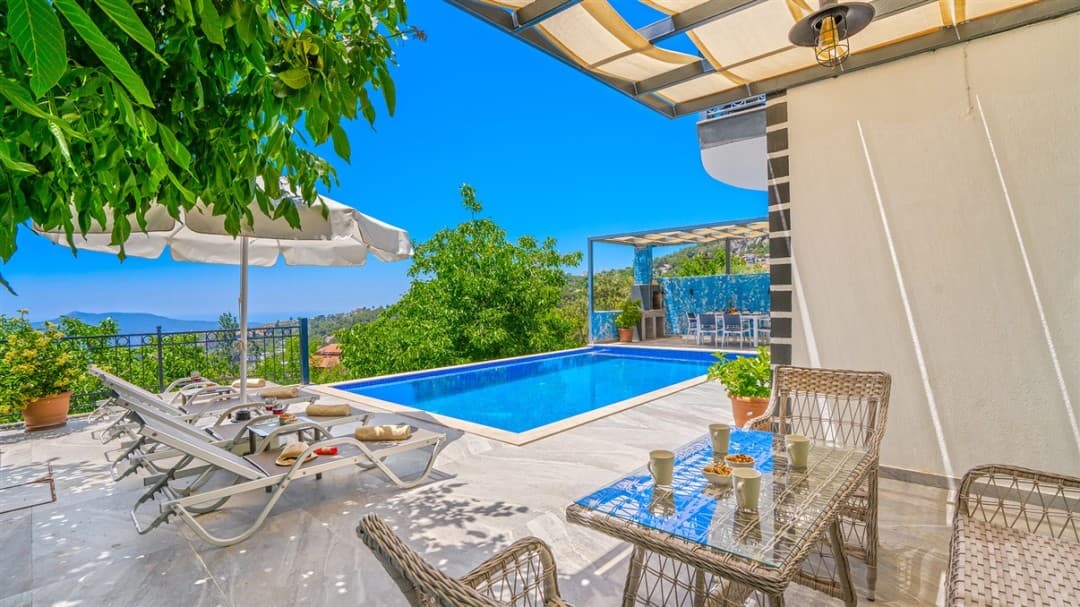 Mavi yüzme havuzu, şezlonglar, güneş şemsiyesi ve bahçe mobilyaları bulunan güneşli bir villa terası.