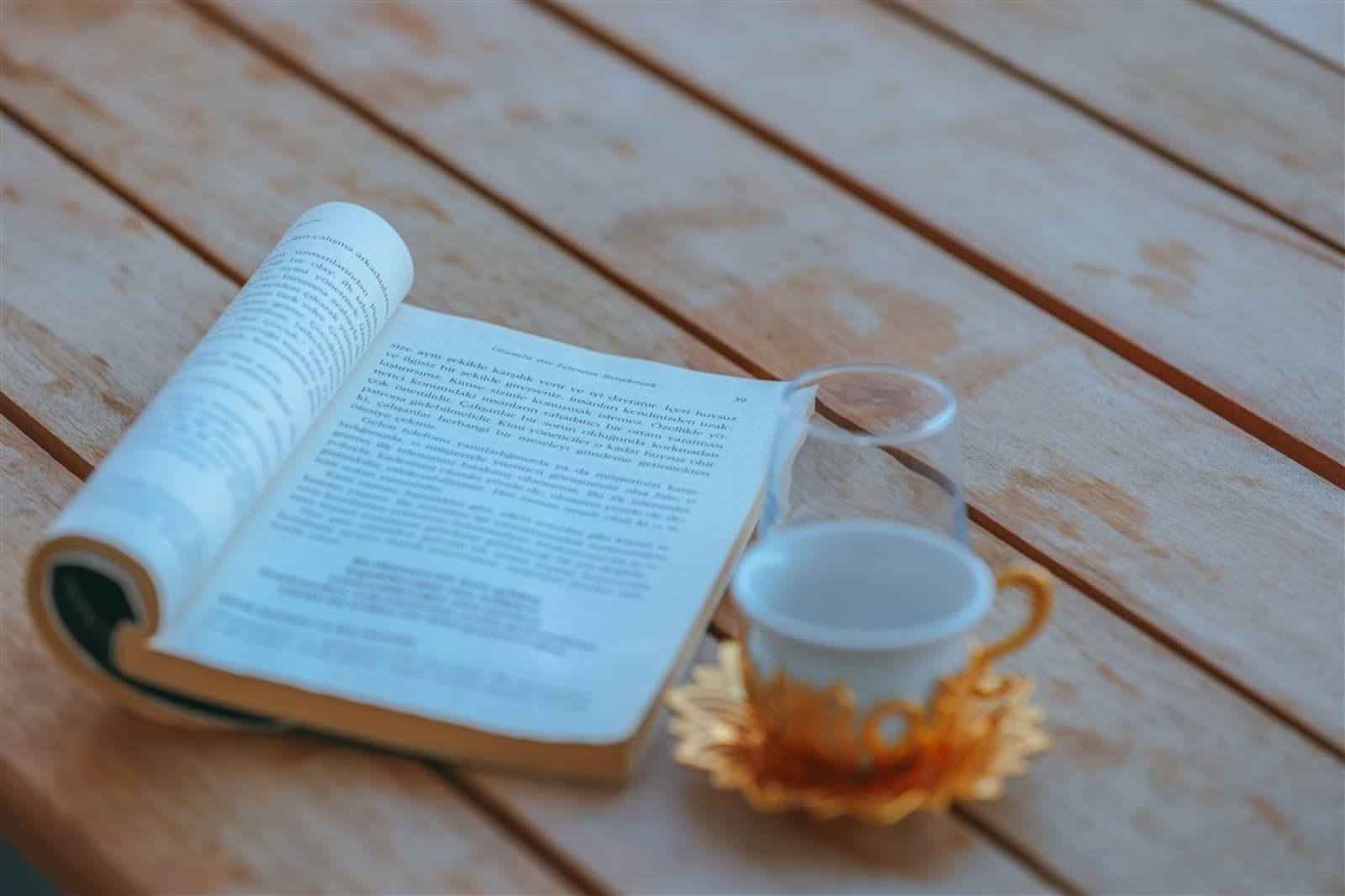 Ahşap yüzey üzerinde açık bir kitap, cam bardak ve fincan tabağı.