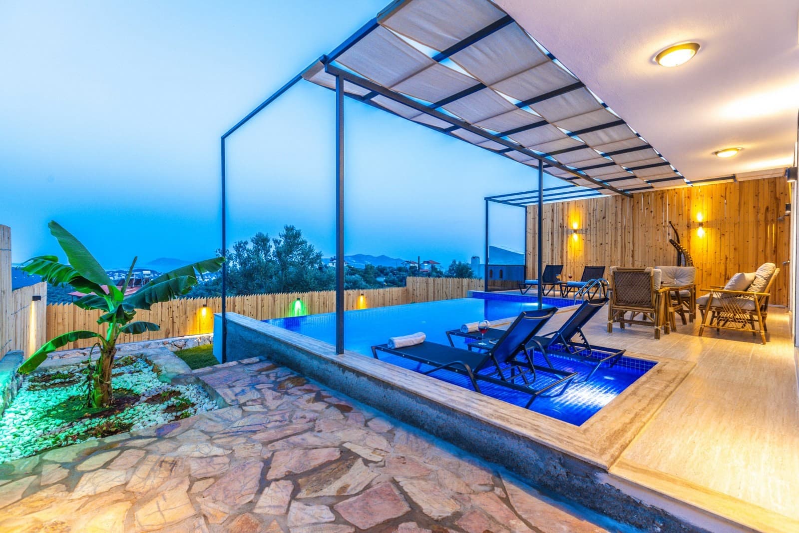Akşam aydınlatmalı, havuz içinde şezlongları ve gölgelikli terası olan manzaralı villa havuzu.