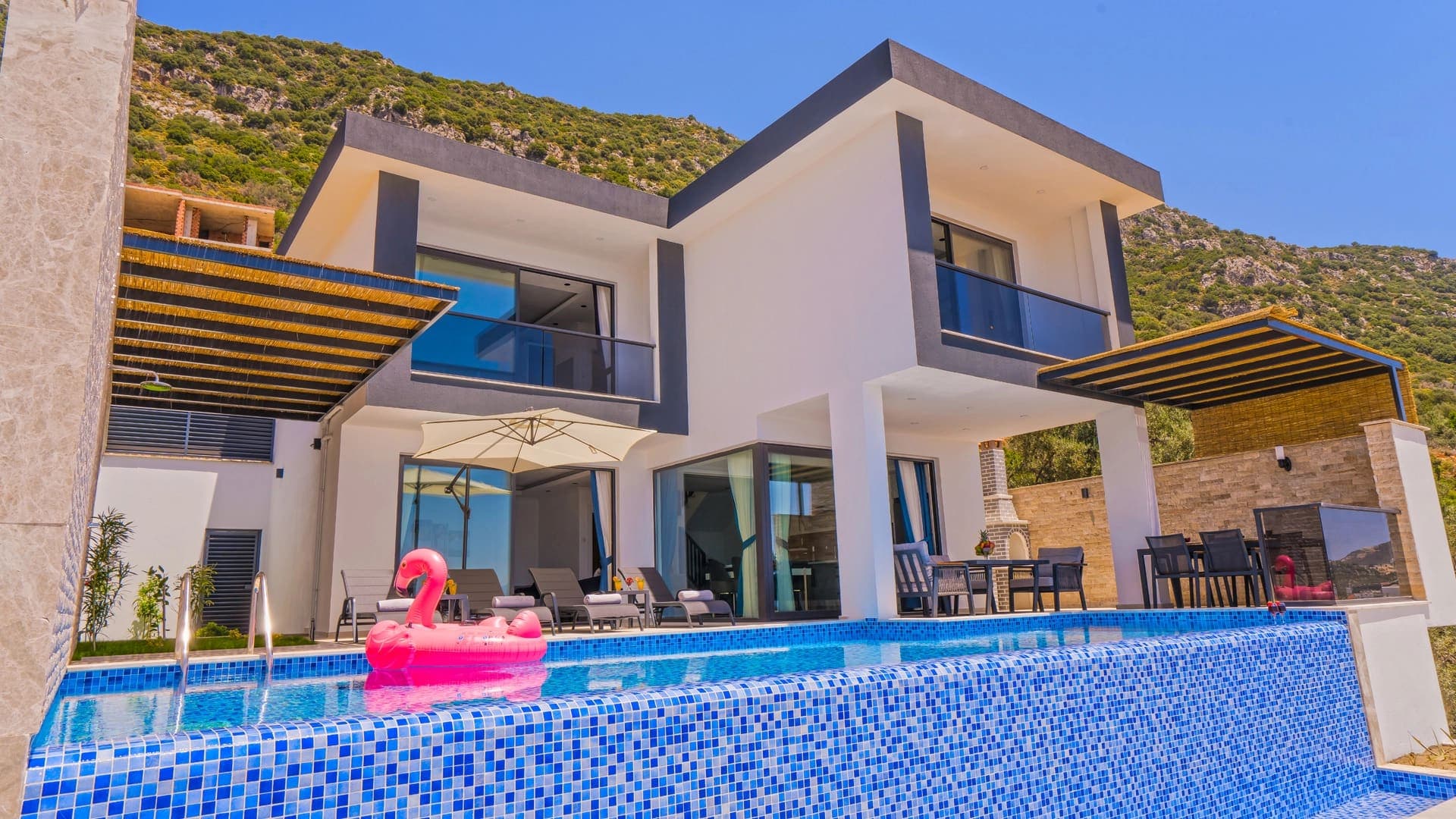 Modern dış cepheli villanın önünde mavi mozaik kaplı havuz, şezlonglar ve pembe flamingo deniz yatağı.