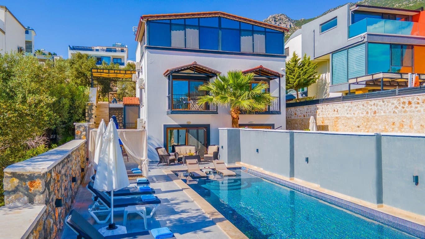 Bahçesinde havuz, şezlonglar ve şemsiyeler bulunan beyaz dış cepheli villa.