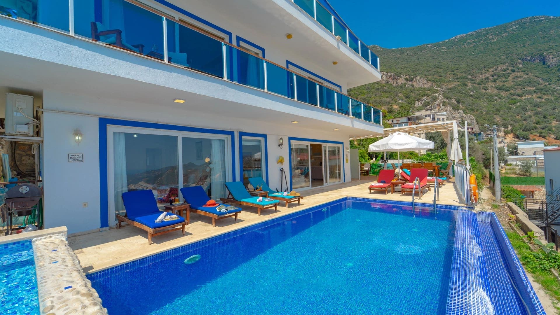 Mavi çerçeveli beyaz villa, havuz kenarında şezlonglar ve güneş şemsiyesi.