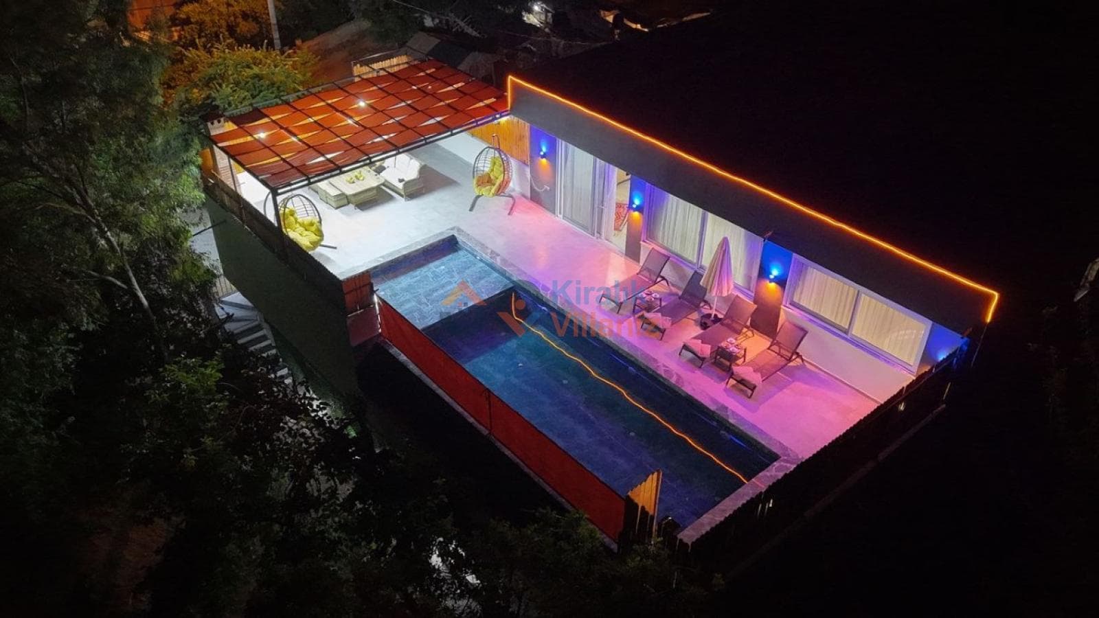 Gece aydınlatmalı, şezlonglu havuz terası ve bahçe manzaralı villa dış görünümü.