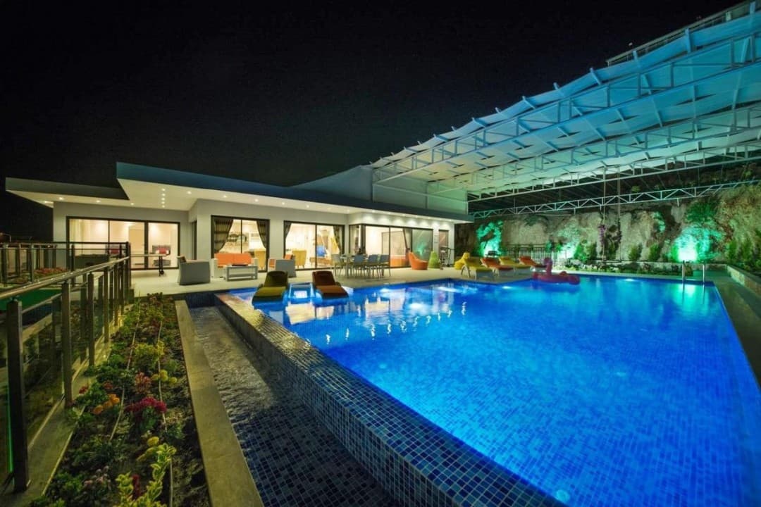 Gece aydınlatmalı büyük yüzme havuzu ve şezlongların bulunduğu villanın dış görünümü.