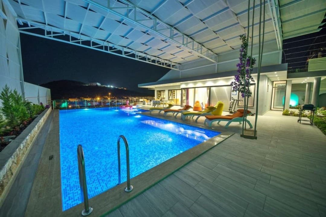 Aydınlatmalı büyük yüzme havuzu, şezlonglar ve gece manzaralı havuz başı terası.