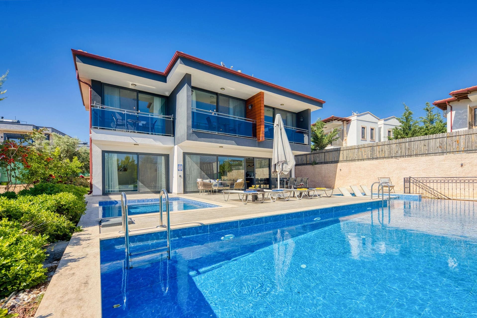 Mavi sığ havuzlu ve şezlonglu villa bahçesi.