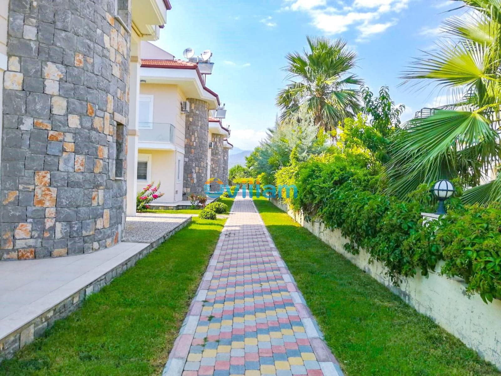 Villa bahçesinde taş duvarlı binanın yanından geçen renkli parke taşlı yürüyüş yolu ve palmiye ağacı.