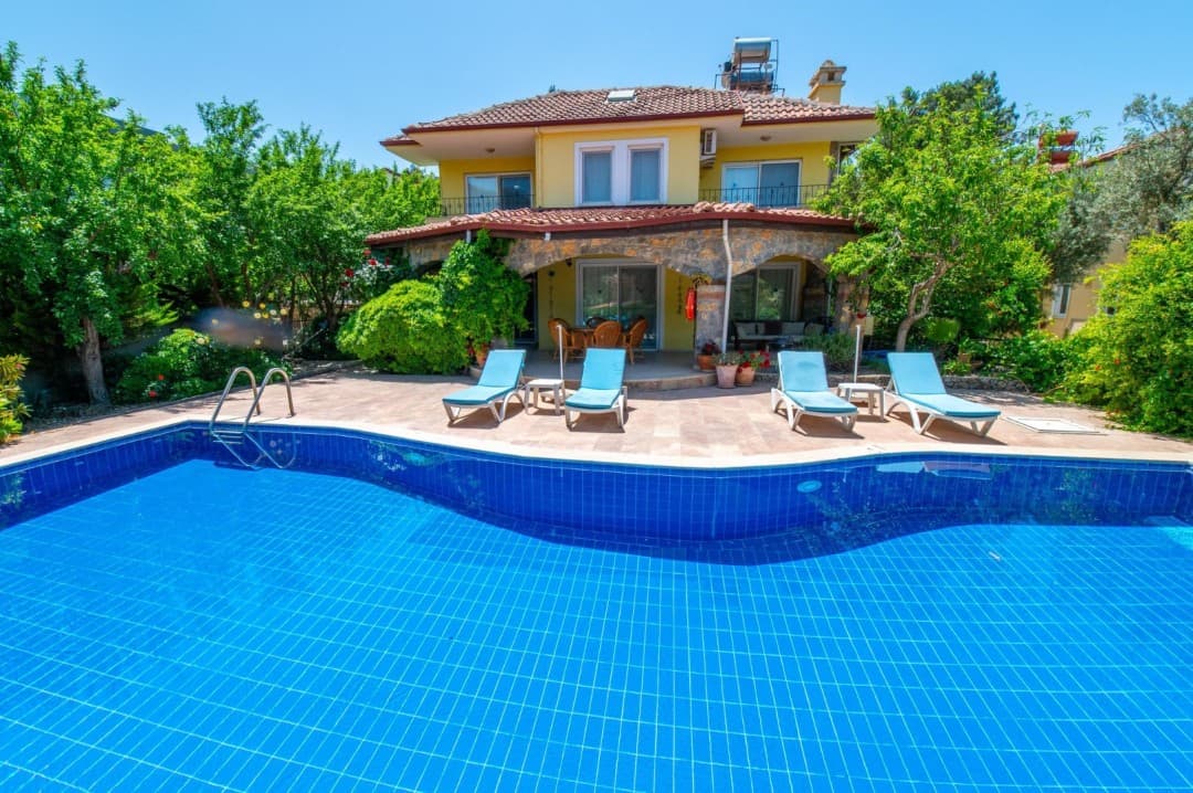 Mavi yüzme havuzu ve şezlongların bulunduğu bahçede sarı renkli iki katlı villa.