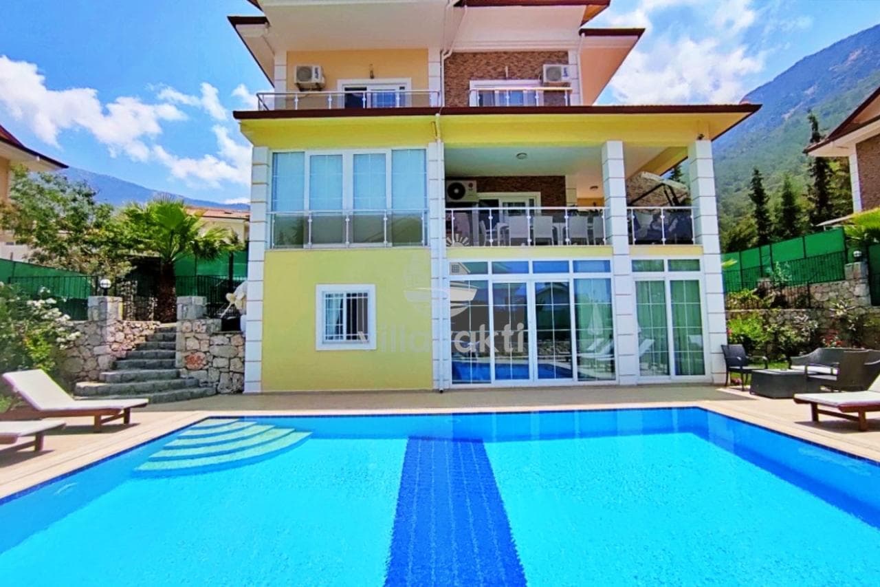 Önünde havuz bulunan, iki katlı, sarı dış cepheli villa ve dağ manzarası.