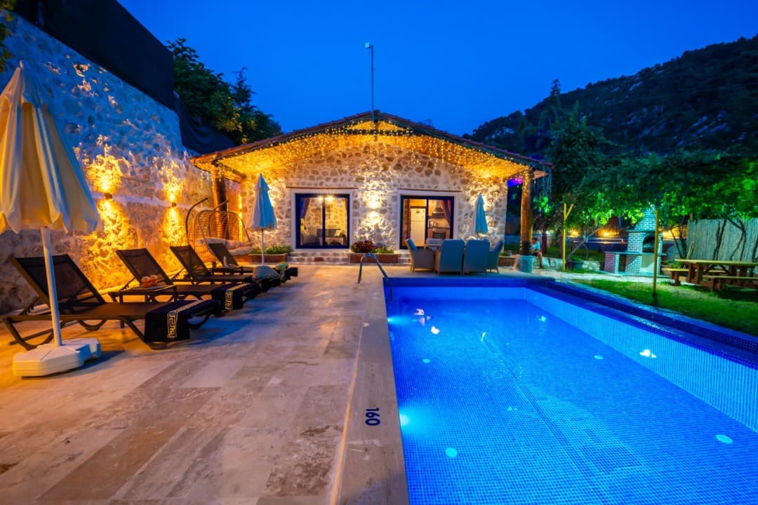 Akşam vakti aydınlatmalı havuz ve şezlongların bulunduğu taş villa.