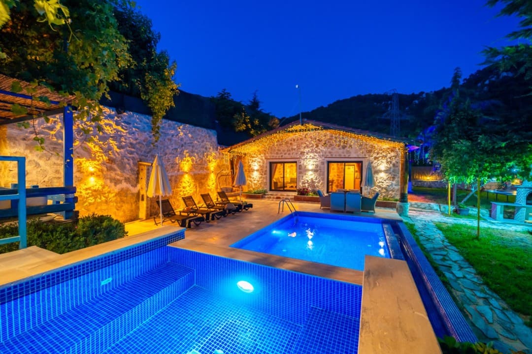 Gece aydınlatmalı taş duvarlı villa, önünde iki kademeli havuz, şezlonglar ve bahçe oturma grubu bulunuyor.