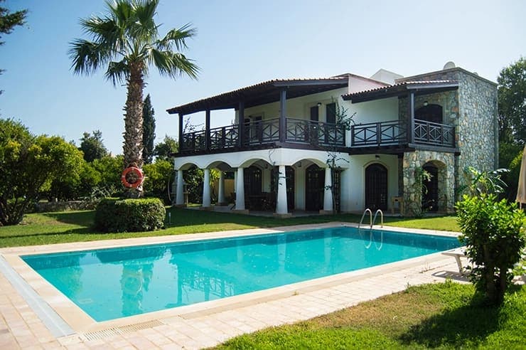 Taş kaplamalı iki katlı villanın önünde palmiye ağacı ve güneşlenme alanı olan açık yüzme havuzu var.