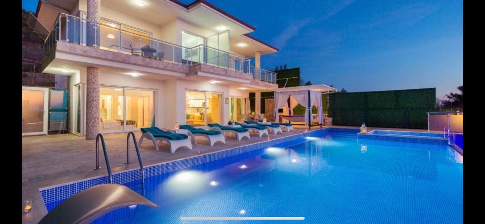 Akşam ışıklandırmalı havuz kenarında şezlonglar, gölgelikli oturma alanı ve modern villa cephesi var.