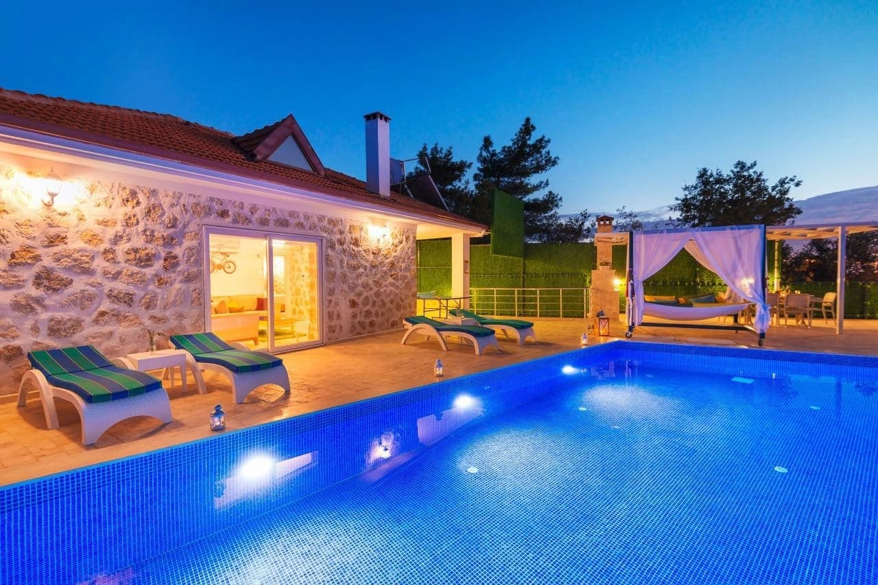 Akşam ışıklandırmalı havuzu ve şezlongları olan taş cepheli villa.
