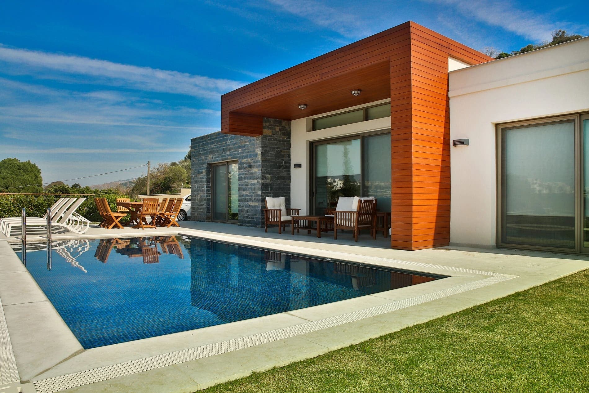 Ahşap kaplamalı modern villa cephesi, önünde mavi yüzme havuzu, şezlonglar ve bahçe mobilyaları var.