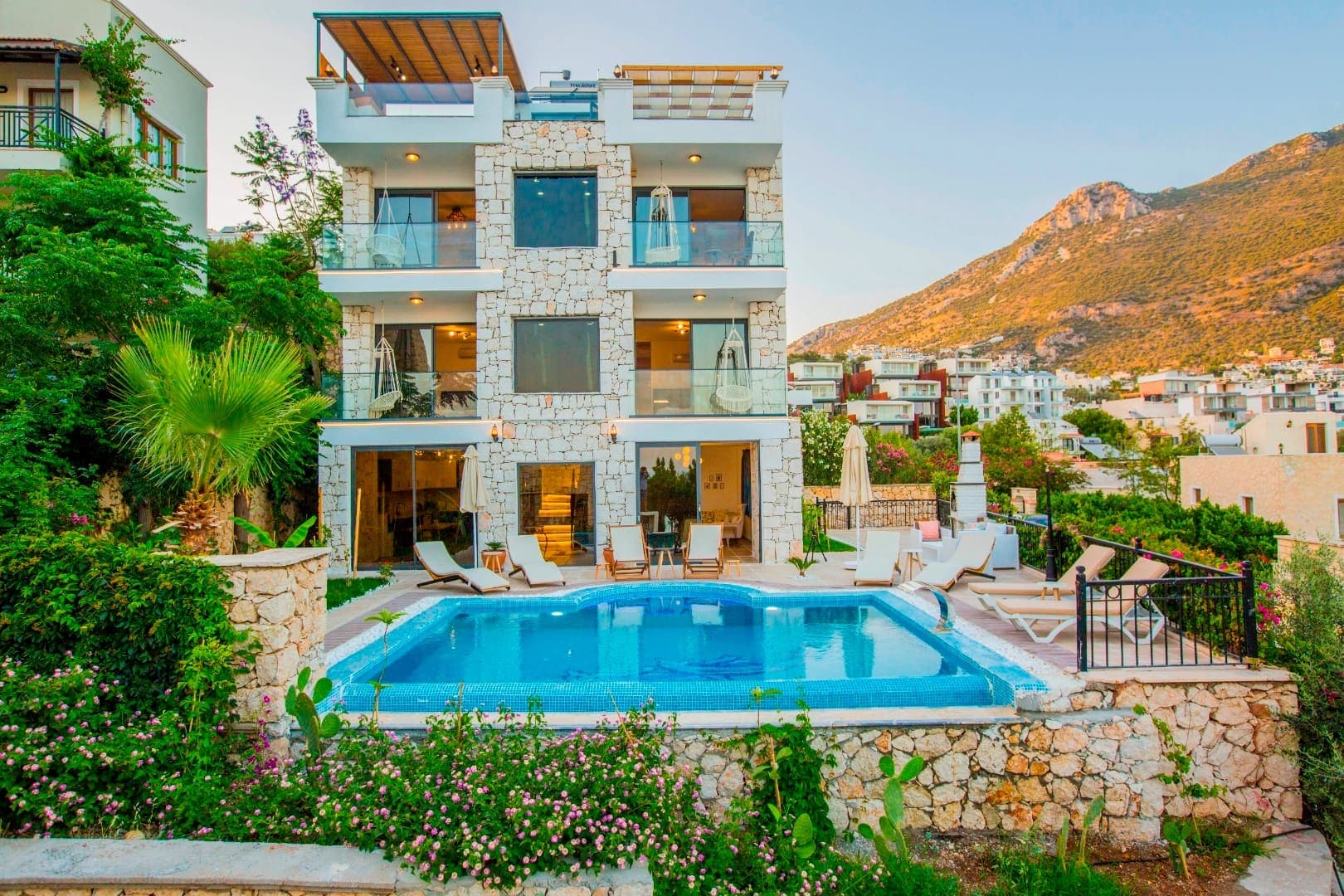 Taş kaplamalı çok katlı villa, önünde yüzme havuzu, şezlonglar ve dağ manzarası bulunuyor.